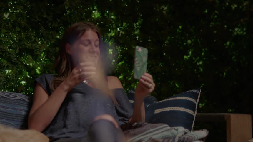 A woman laughs and points while having a drink and face timing on her phone sitting outside by the fire