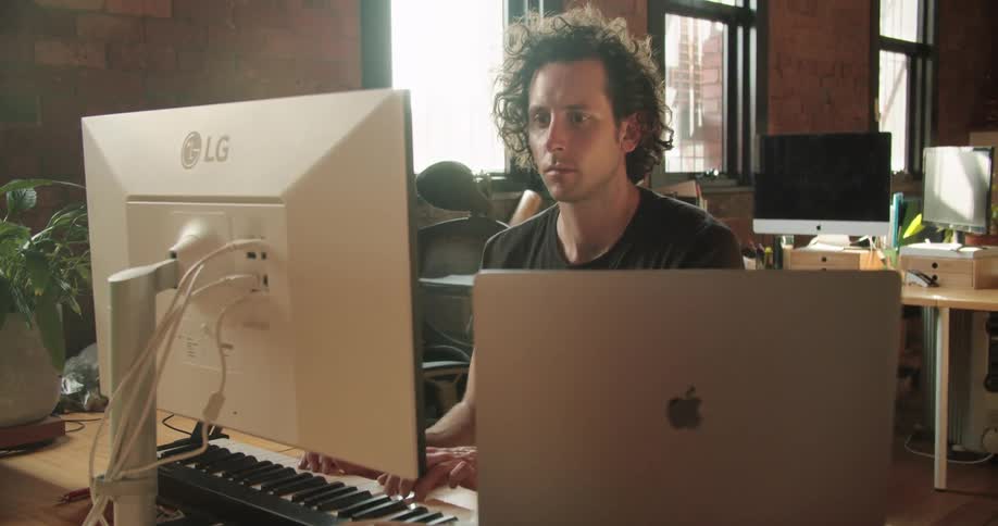 A man with curly hair plays an electronic keyboard connected to a computer while looking at a computer monitor