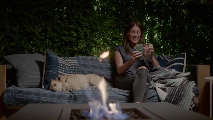 Woman sits on outdoor couch near fire pit with sleeping dog