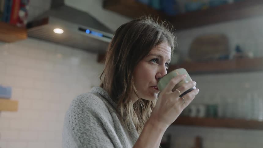 A woman drinks from a coffee mug standing in a kitchen