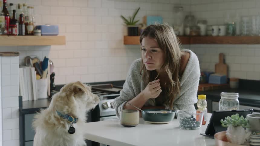 A woman sitting at her dining table eats food from a bowl and pets her dog