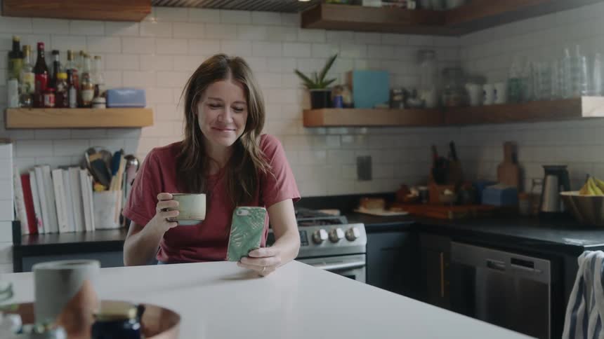 A woman sits at her kitchen table holding a coffee cup and having a video conversation on her phone