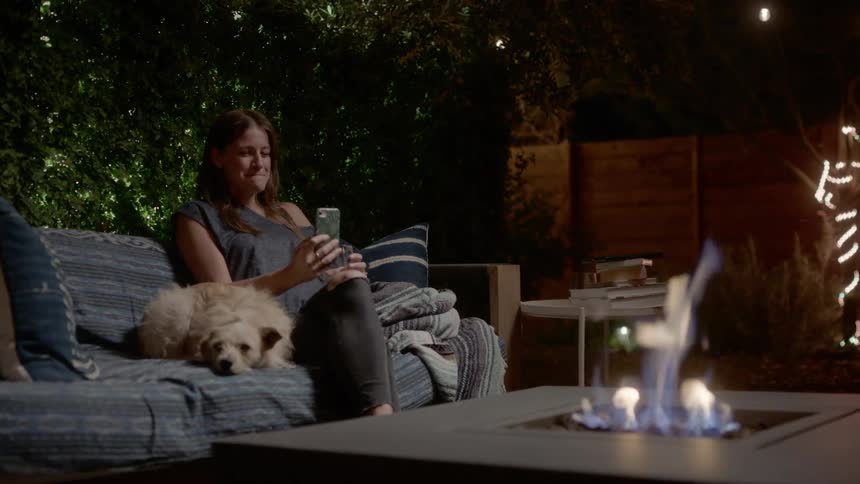 Woman talks into her phone while sitting on a couch next to her dog outside near a fire