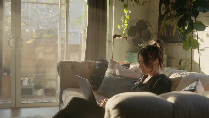 Woman sits on sofa in sunny room using laptop
