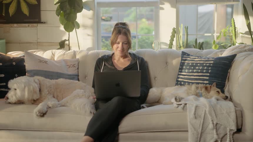 Woman sits on the sofa between two white dogs as she works on a laptop