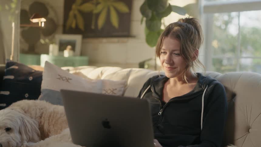 A woman smiles while using a laptop on a couch at home