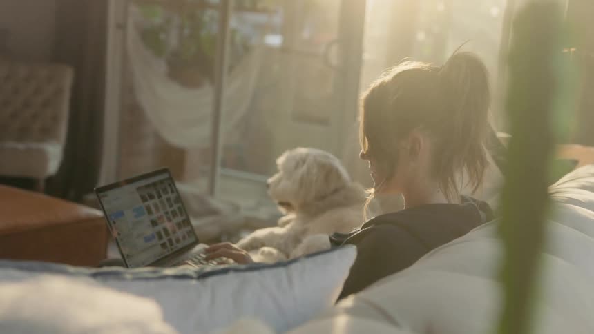 A woman sits on the couch with her dog while using her laptop