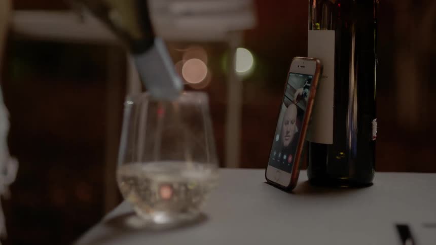 A person pours a glass of wine while video chatting with someone on their phone