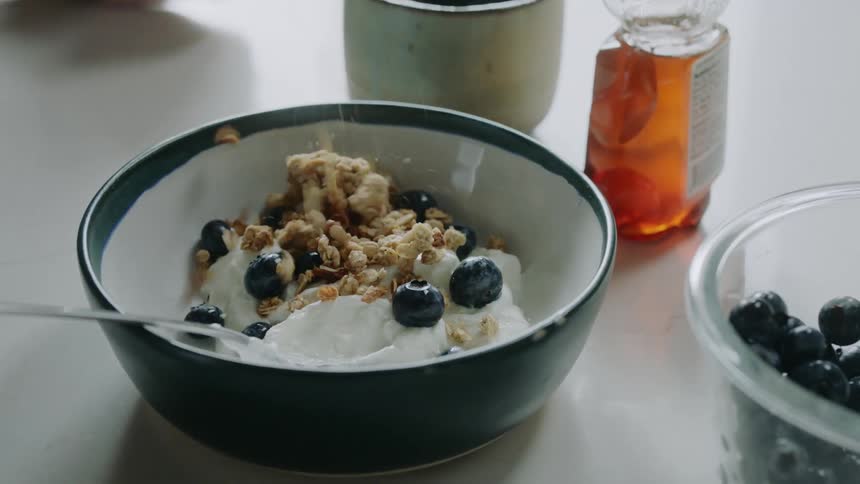 A person pours granola on top of a bowl of yogurt and blueberries