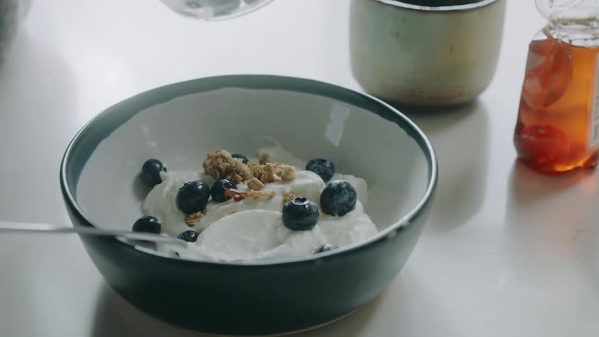 A woman adds blueberries to a bowl of yogurt with a spoon in it