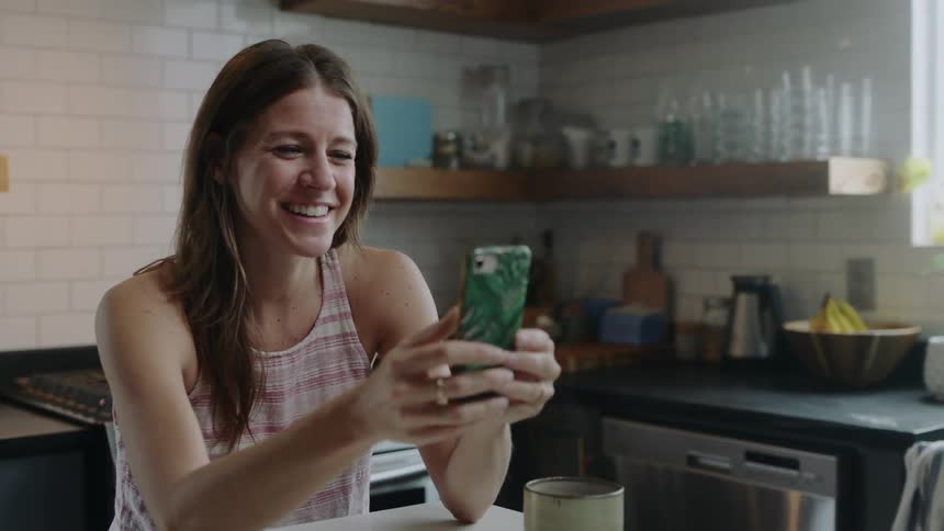 Female standing with her elbows on a kitchen island with a steaming cup of coffee is laughing while having a video conversation on her cellphone