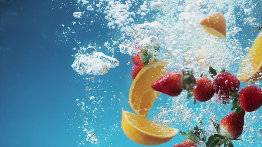 A whole lime along with orange sections and strawberries splash down through water causing bubbles to rise against a blue background