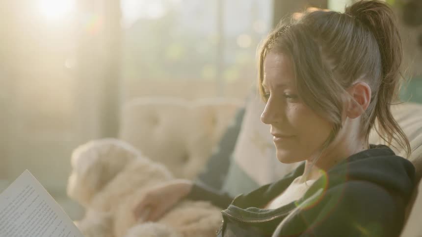 A woman sits on a couch reading a book and petting a dog