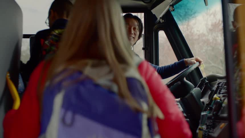 A driver smiles while children climb onto a yellow school bus