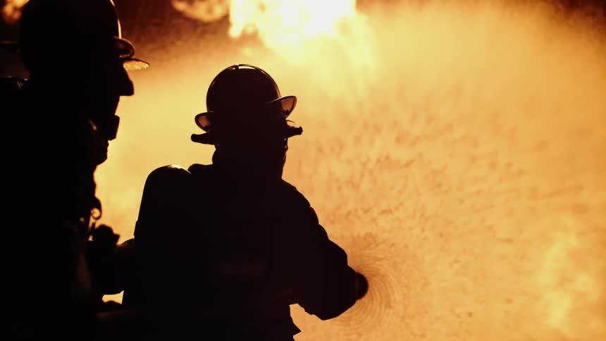 Firefighters backlit by flames spraying hose on fire