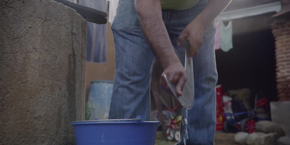 Man cleaning a very large knife with water from a blue bucket