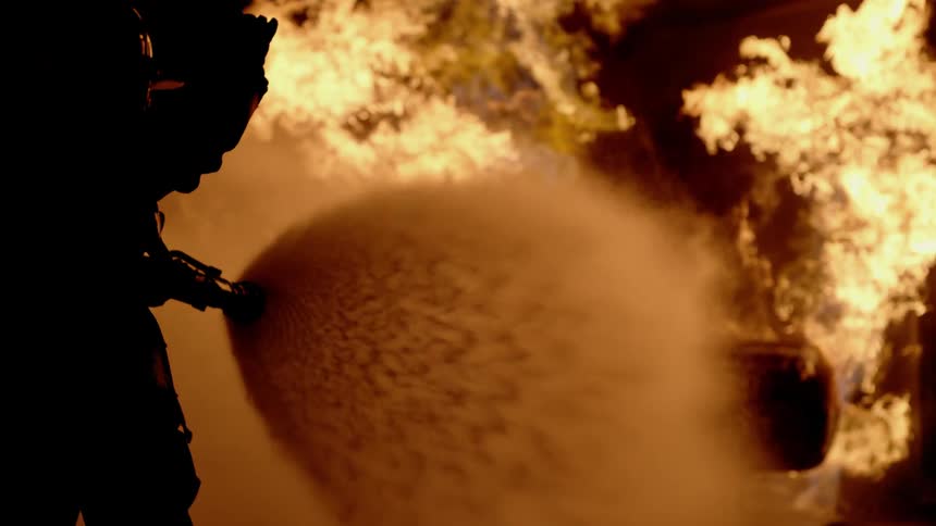 A firefighter sprays water on to a large raging fire in slow motion