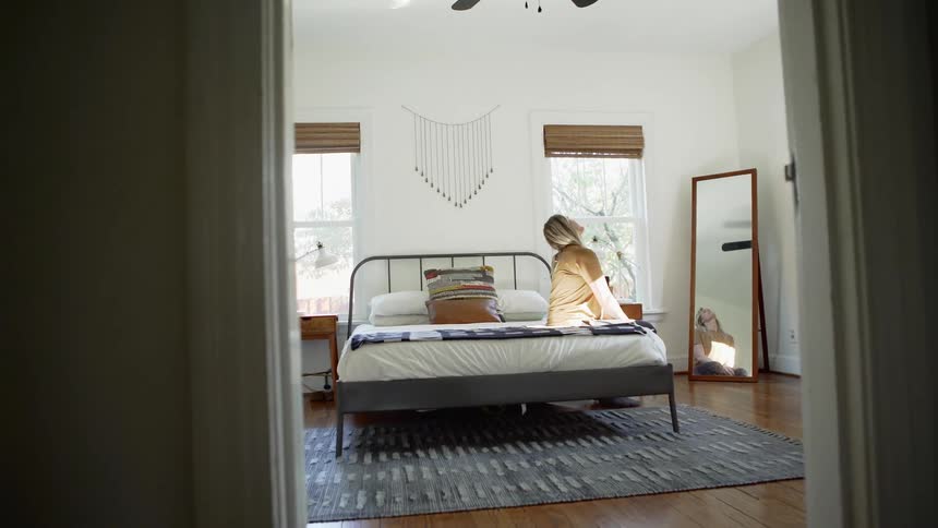 A woman sits on the edge of her bed and stretches her neck