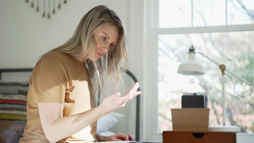 A woman sits on the edge of her bed looking down with her hands in her lap