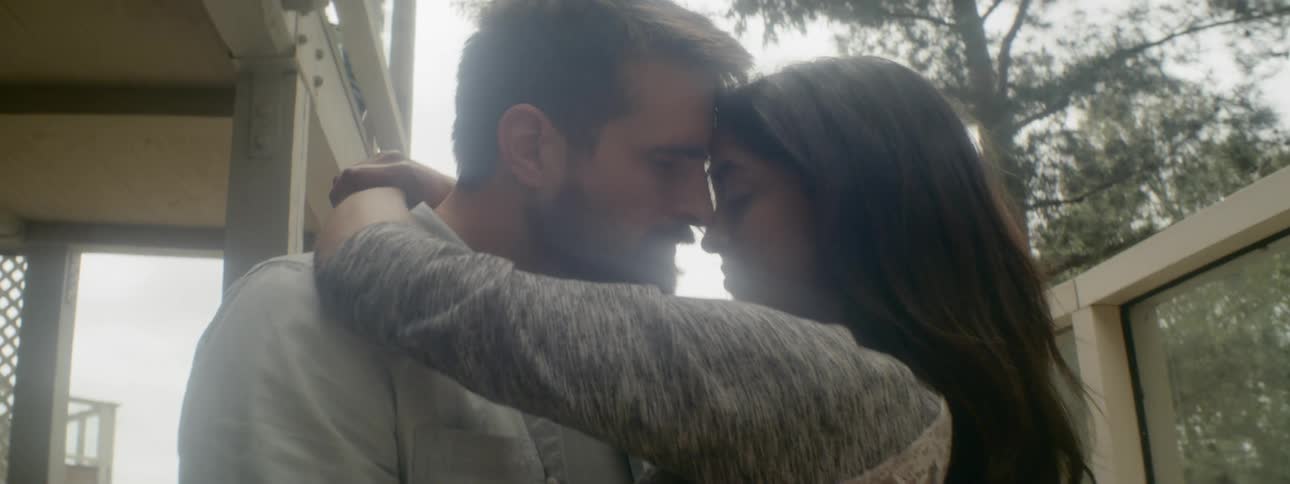 A man and woman stand face to face embracing and caressing each other outside in their yard