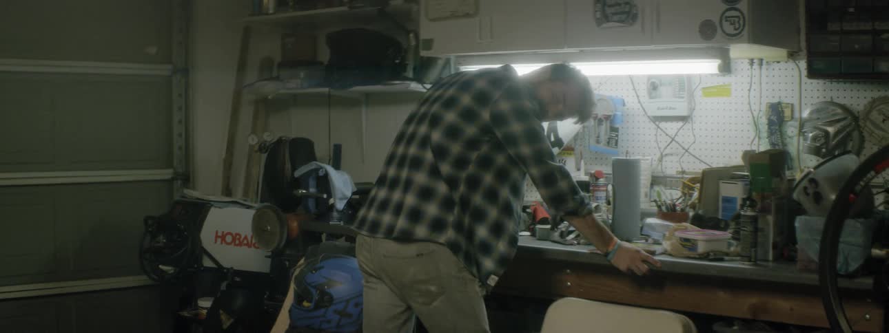 A man in a plaid shirt leans over a workbench in a garage