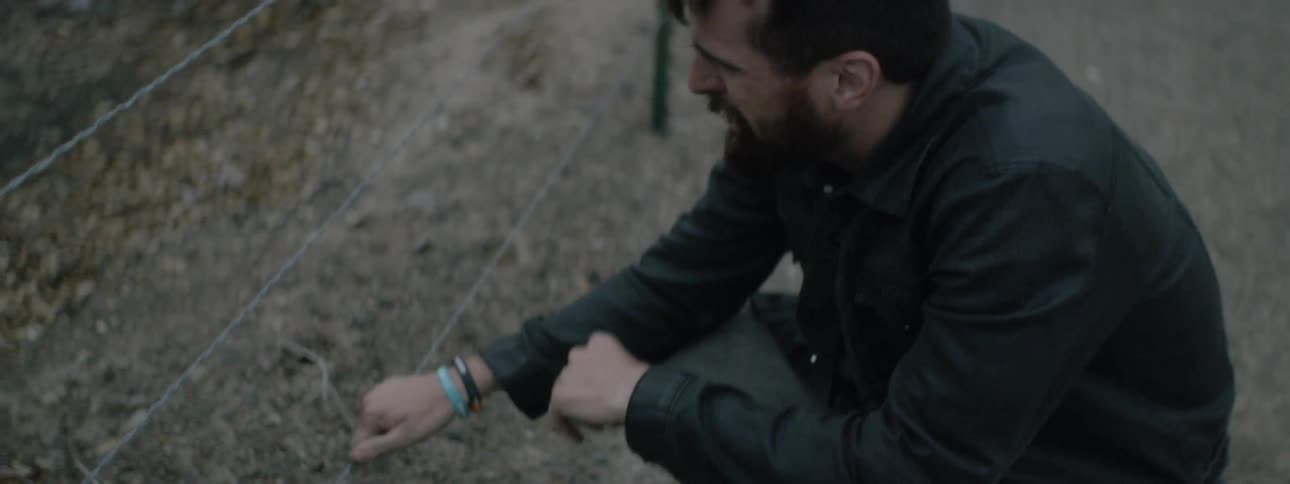 Man crouches down near side of road holding wire fence and crying