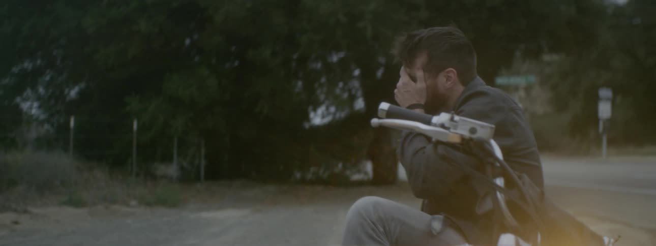 A man sits on his motorcycle with his hand up to his face thinking and looking upset
