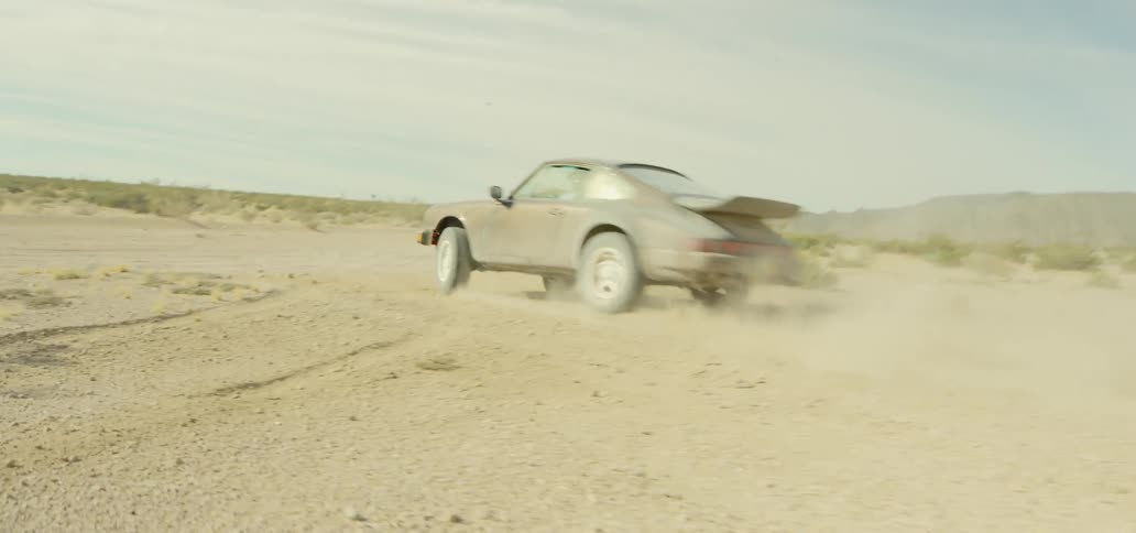 A porsche sports car speeds through dirt filled off road area before making a hairpin turn
