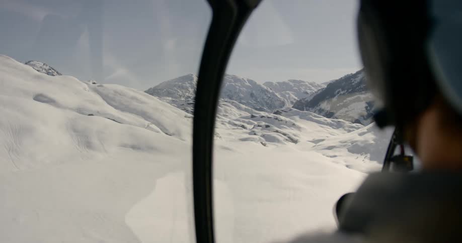 A pilot flies a helicopter low over a rocky snow covered mountains slope on a sunny day