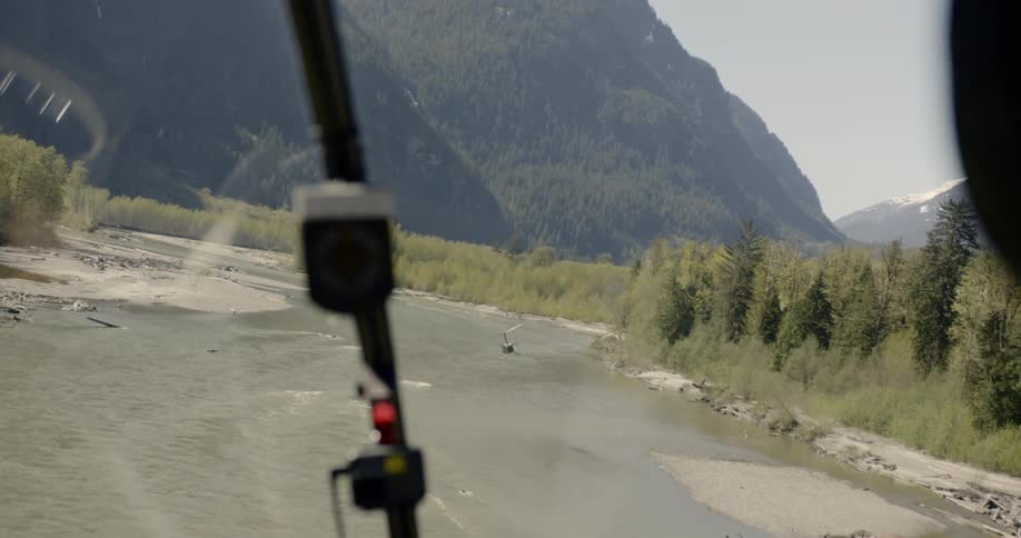 One helicopter follows another flying over river toward a mountain