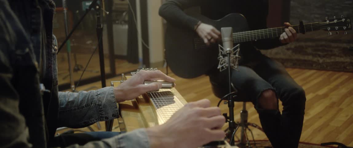 Two male musicians sit in a studio while one plays an acoustic guitar and the other plays a steel guitar