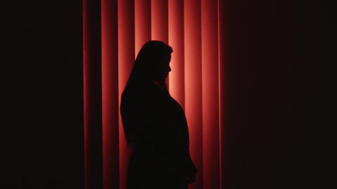 The silhouette of a woman standing in front of blinds that are shaded red from a light