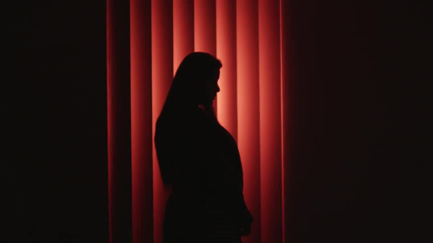 The silhouette of a woman standing in front of blinds that are shaded red from a light
