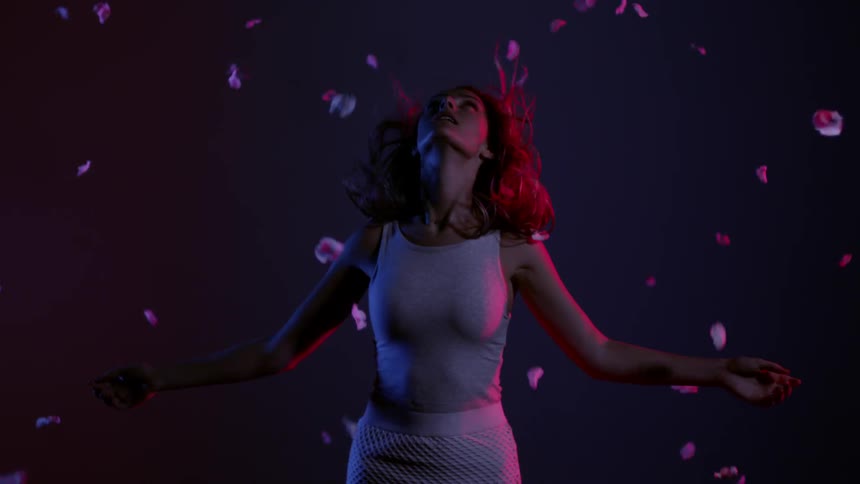 A woman standing against a gray background as her hair blows around her face and rose petals fall around her