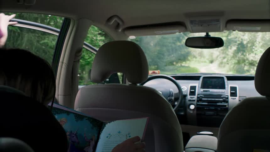 A woman gets into the front seat of her vehicle as her child reads a book in the backseat