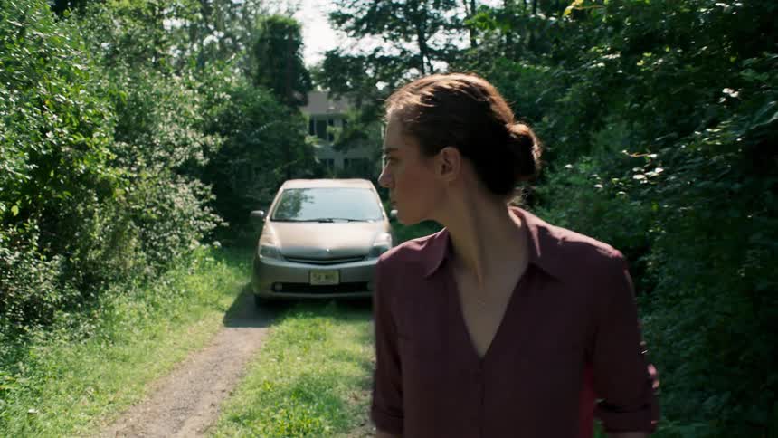 A woman walks along a dirt road in a forested area with her car behind her