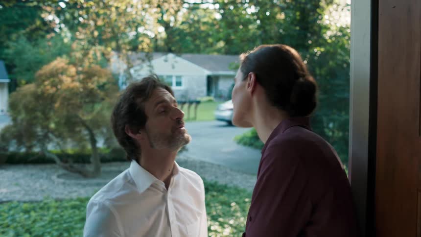 A woman stands at the doorway and greets her husband with a kiss as he and their daughter enter the house