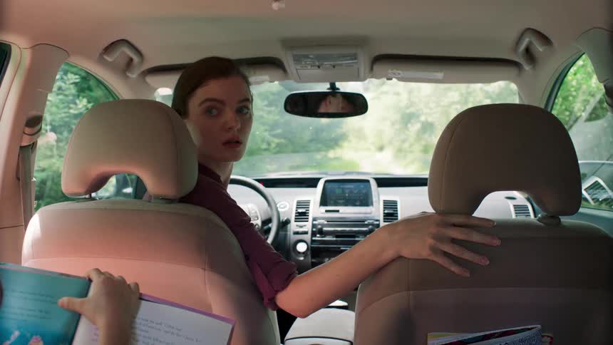 A woman backs her vehicle out of the driveway as her child reads a book in the backseat