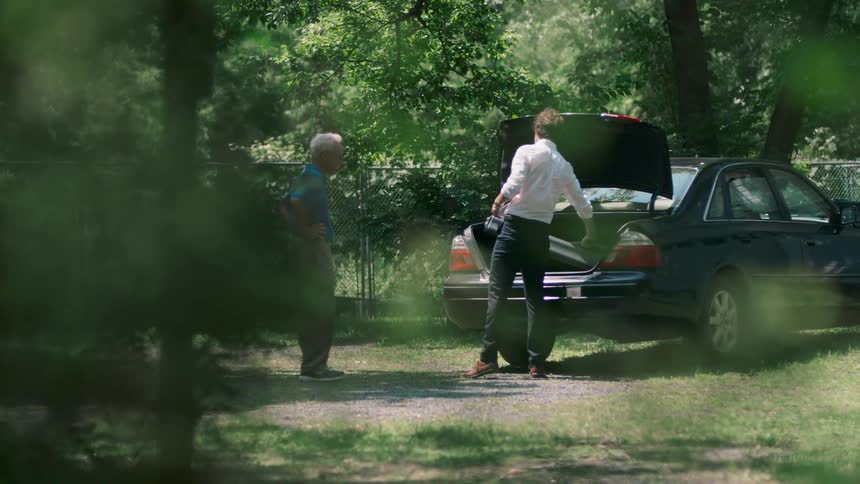 A pair of men standing together in a woodland while one places a case in the trunk of a vehicle