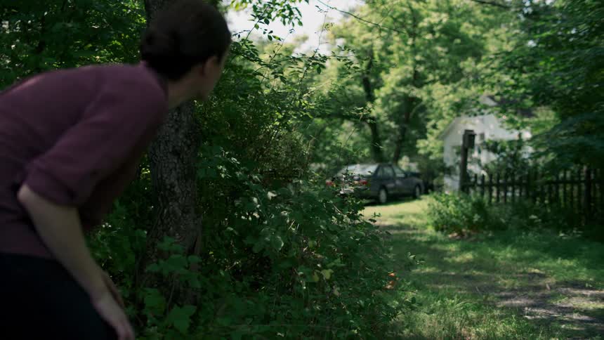A woman hides behind a tree and looks toward a home