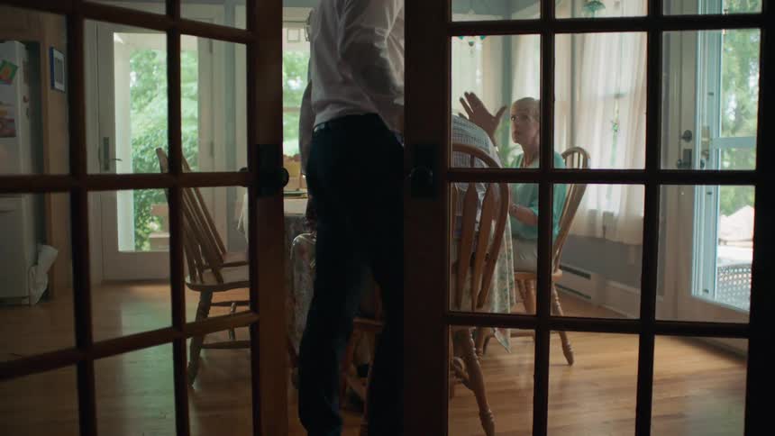 A man enters the dining room where his parents are seated through french doors carrying a wine glass