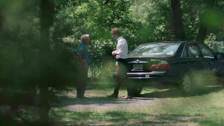 A man closes his car trunk while chatting with another man in his driveway