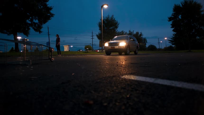 A person stands near a car with headlights on in an empty parking lot with an overturned cart at night