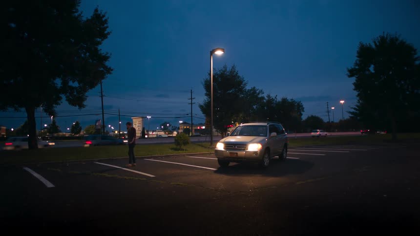 A man talks on a phone while walking around his vehicle in a parking lot