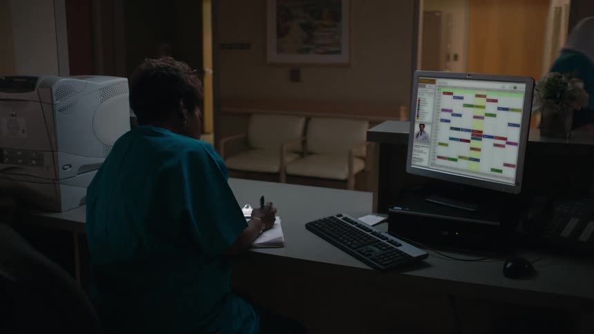 A woman in blue scrubs sits at a desk writing notes while a nurse walks by