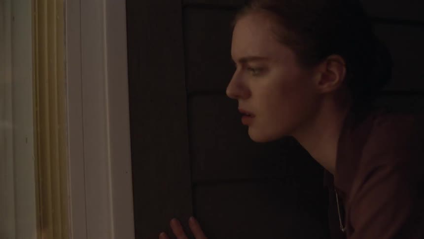 A woman slowly stands as she looks into a house window while trying to remain hidden