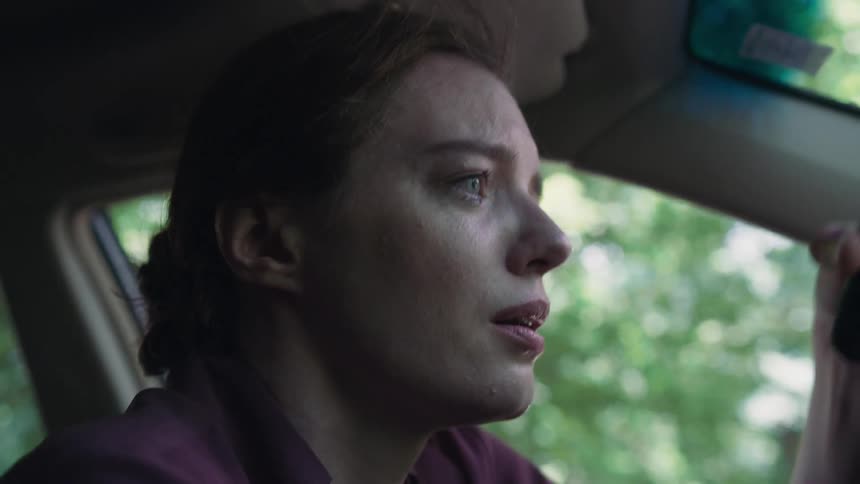 A woman sitting in a car anxiously reaches for a phone on the dashboard