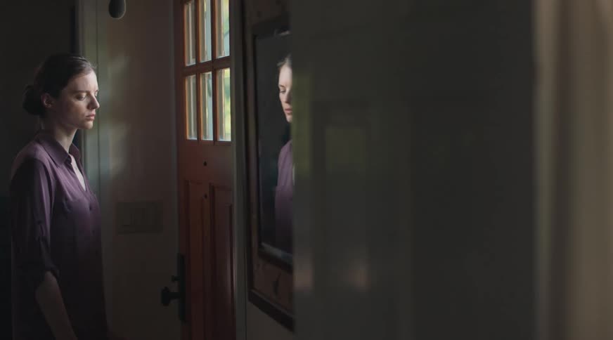 A woman looking very sad looks in a mirror and then walks away