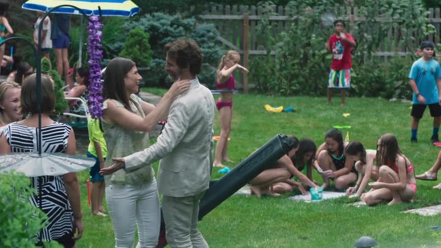 A man carrying a case enters a backyard pool party and greets his children then sneaks up on his wife and kisses her