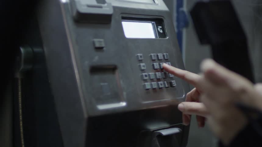 A pair of hands pick up a public phone and begin to dial using the keypad